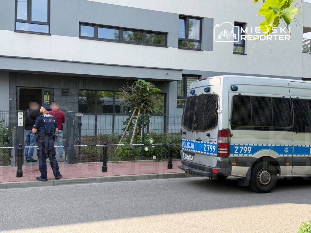 Policjant rozmawia z dwoma mężczyznami przed nowoczesnym budynkiem mieszkalnym, obok stoi radiowóz policyjny.
