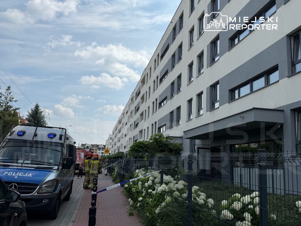 W pobliżu nowoczesnego budynku mieszkalnego grupka strażaków i policjantów pracuje przy akcji ratunkowej.