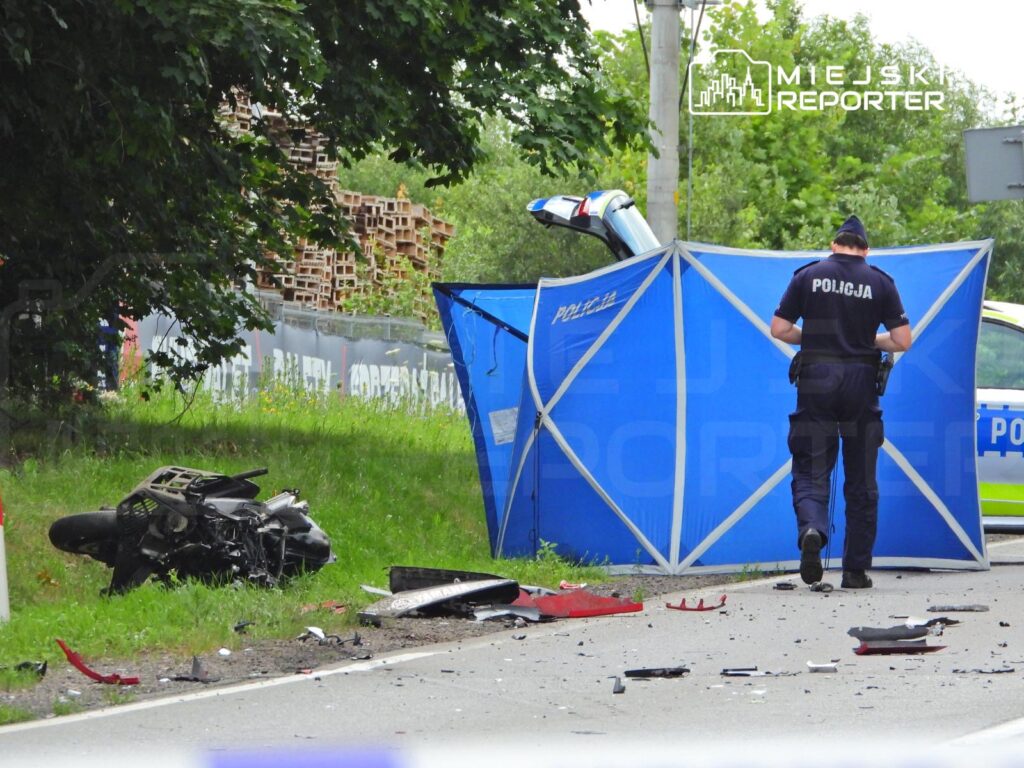 Motocykl leży na poboczu drogi, a policjant bada miejsce wypadku, przy niebieskiej osłonie.