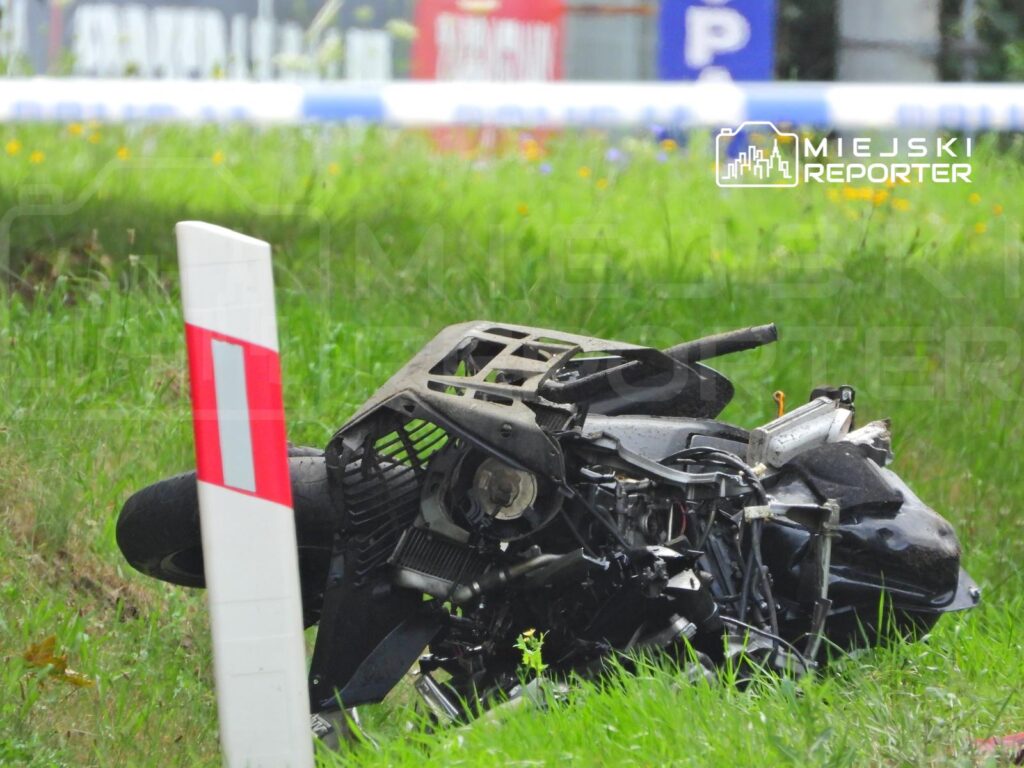 Zniszczony motocykl leżący na trawie obok słupa z oznaczeniem drogowym.
