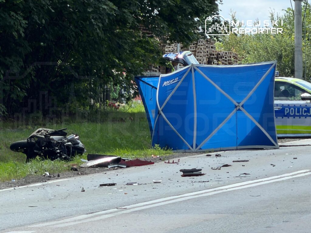 Zniszczony motocykl leży na poboczu drogi obok niebieskiego namiotu policyjnego, a w tle widać radiowóz.