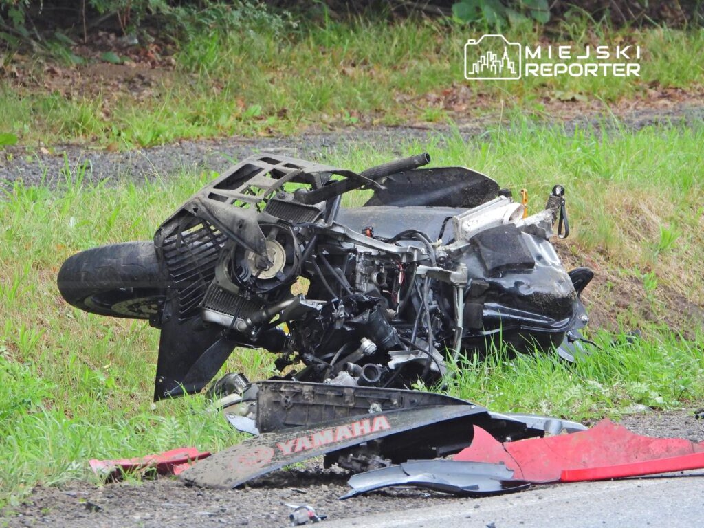 Zniszczony motocykl leży na poboczu drogi, otoczony trawą i fragmentami karoserii.
