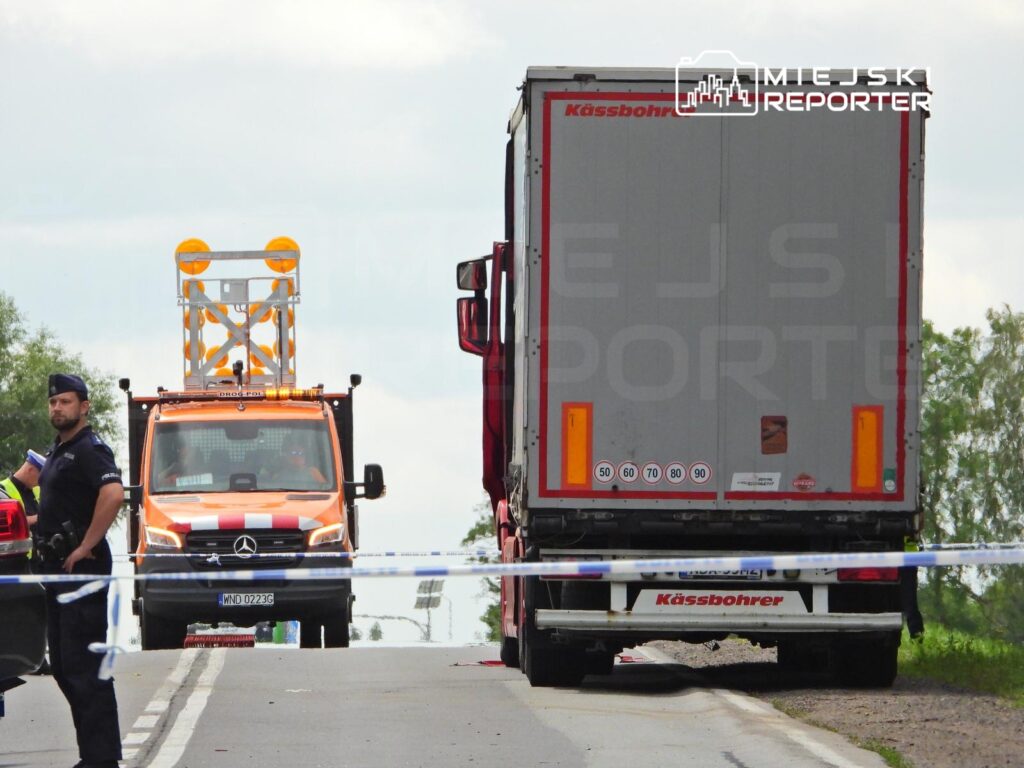 Policjant kierujący ruchem obok ciężarówki na drodze, z samochodem służb ratunkowych w tle.