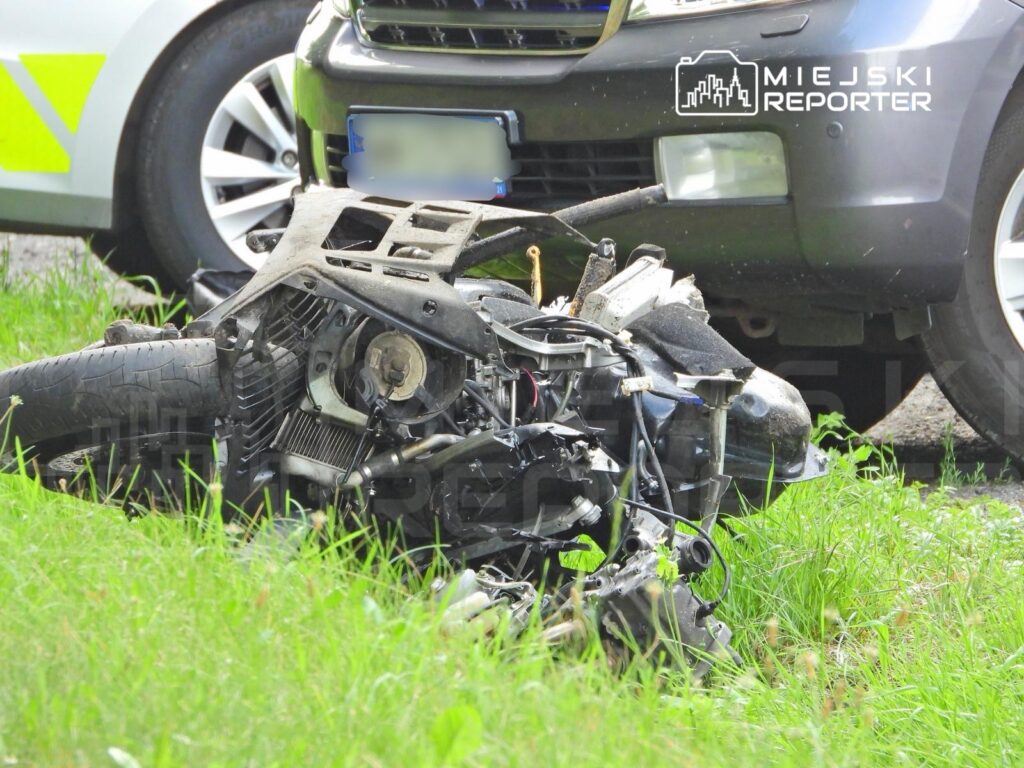 Zniszczony motocykl leży na trawie obok zaparkowanego samochodu.