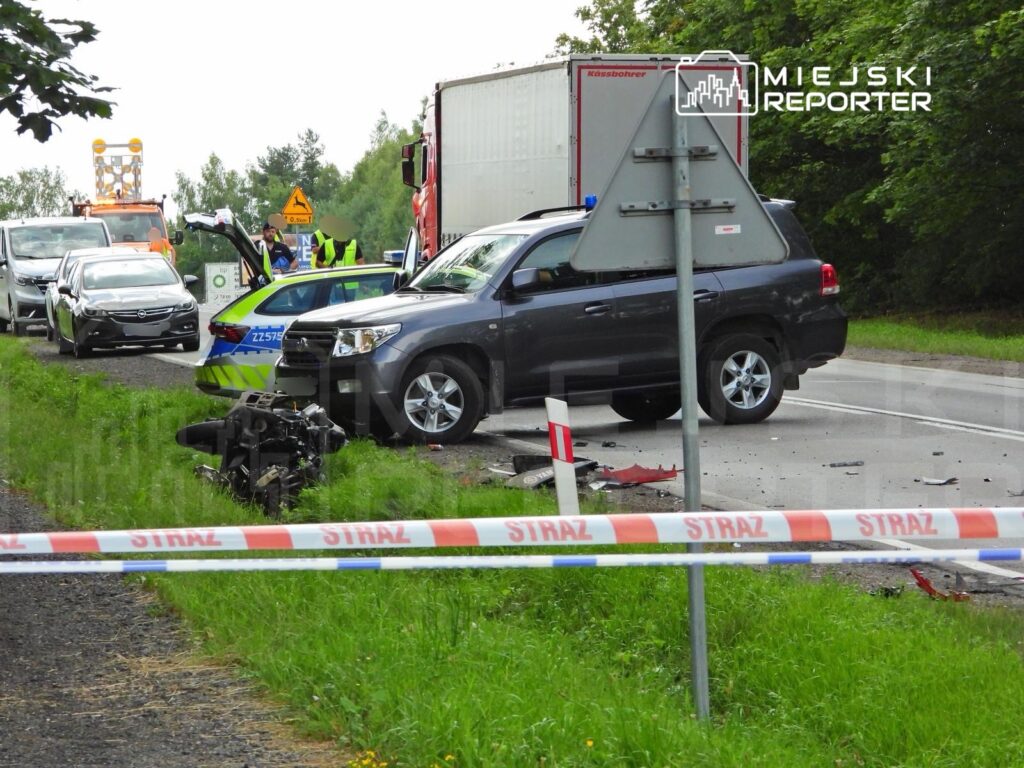 Na drodze widać wypadek samochodowy z uszkodzonymi pojazdami, w tym osobowym i ciężarowym. W tle pracują służby ratunkowe.