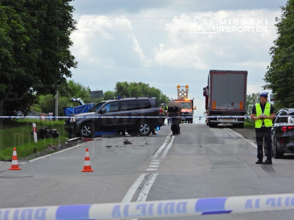 W miejscu zdarzenia drogowego leży uszkodzony samochód osobowy, a obok stoją funkcjonariusze policji w kamizelkach odblaskowych.