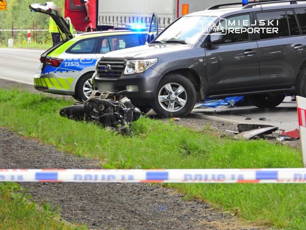 Wypadek drogowy z uszkodzonym silnikiem motocykla na poboczu, obok policyjnego radiowozu i ciężarówki.