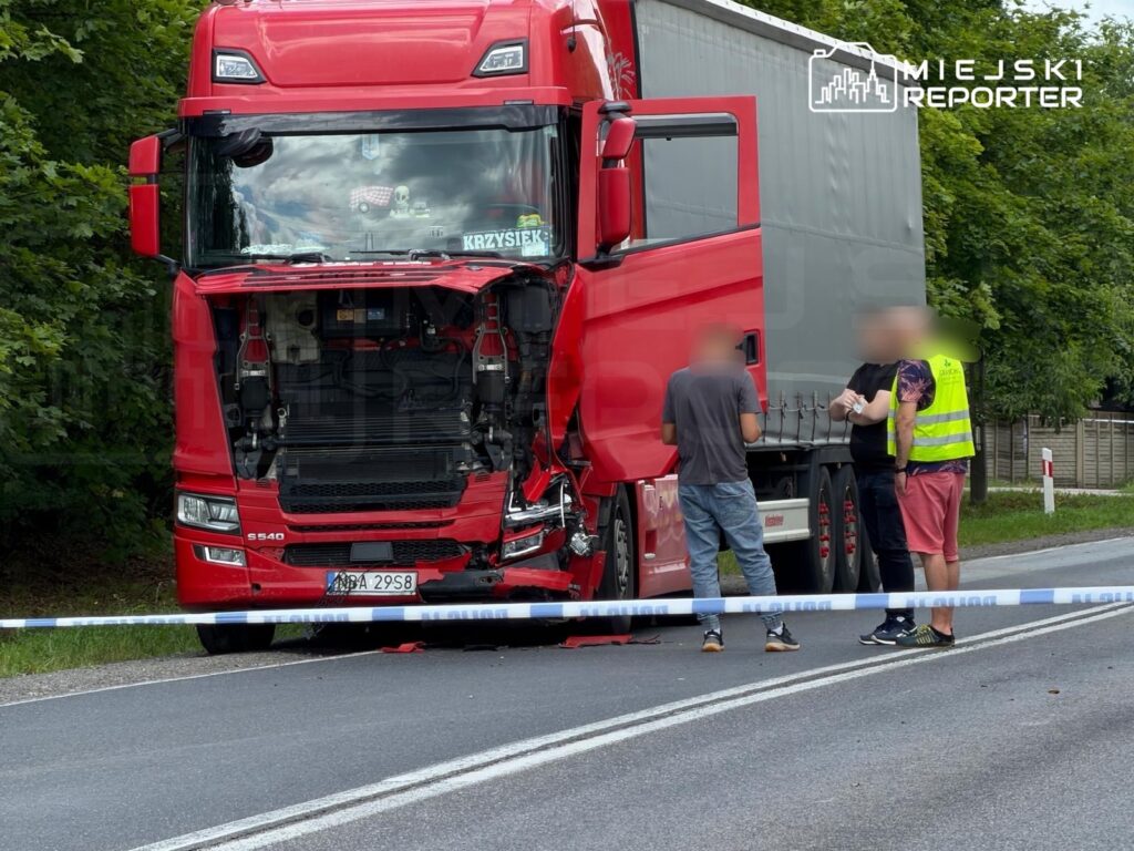 Czerwona ciężarówka z otwartą maską stoi na drodze, a trzech mężczyzn rozmawia obok niej. W tle drzewa i barierka.
