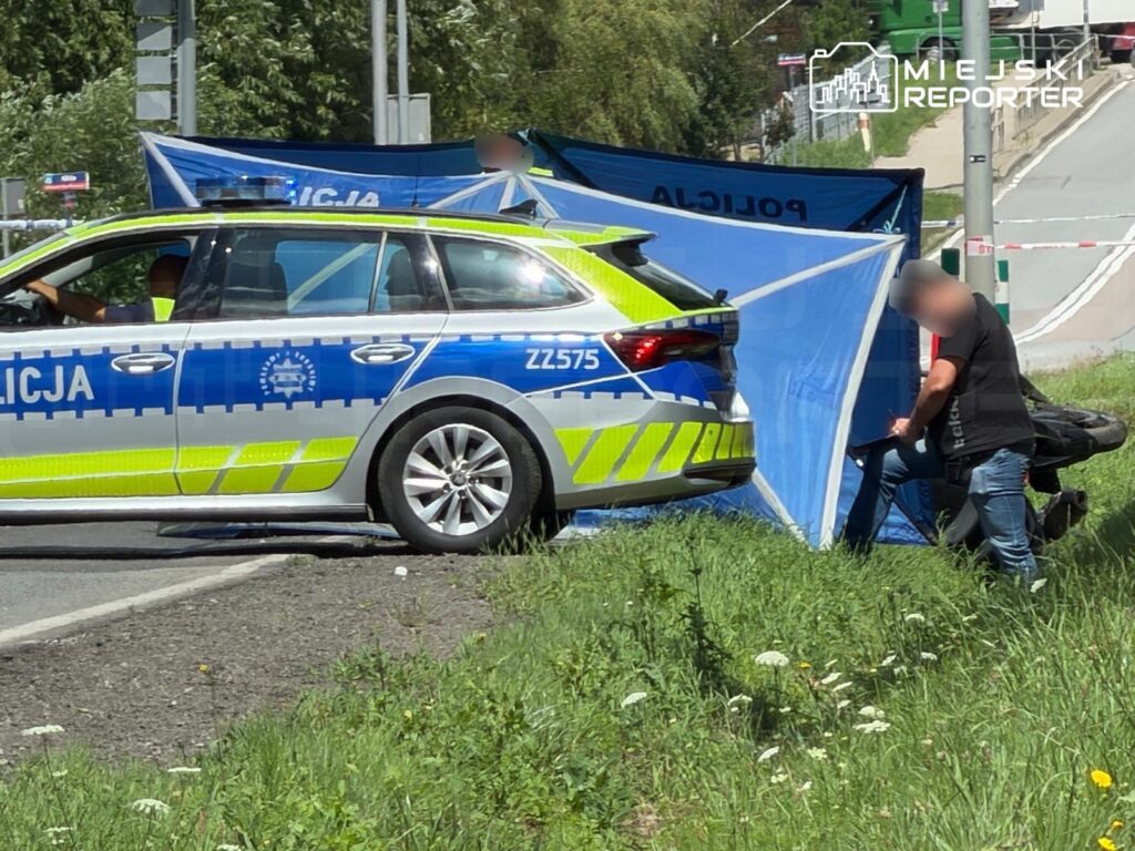 Mężczyzna w czarnej koszulce siedzi na krześle obok policyjnego samochodu, na tle niebieskiego namiotu.