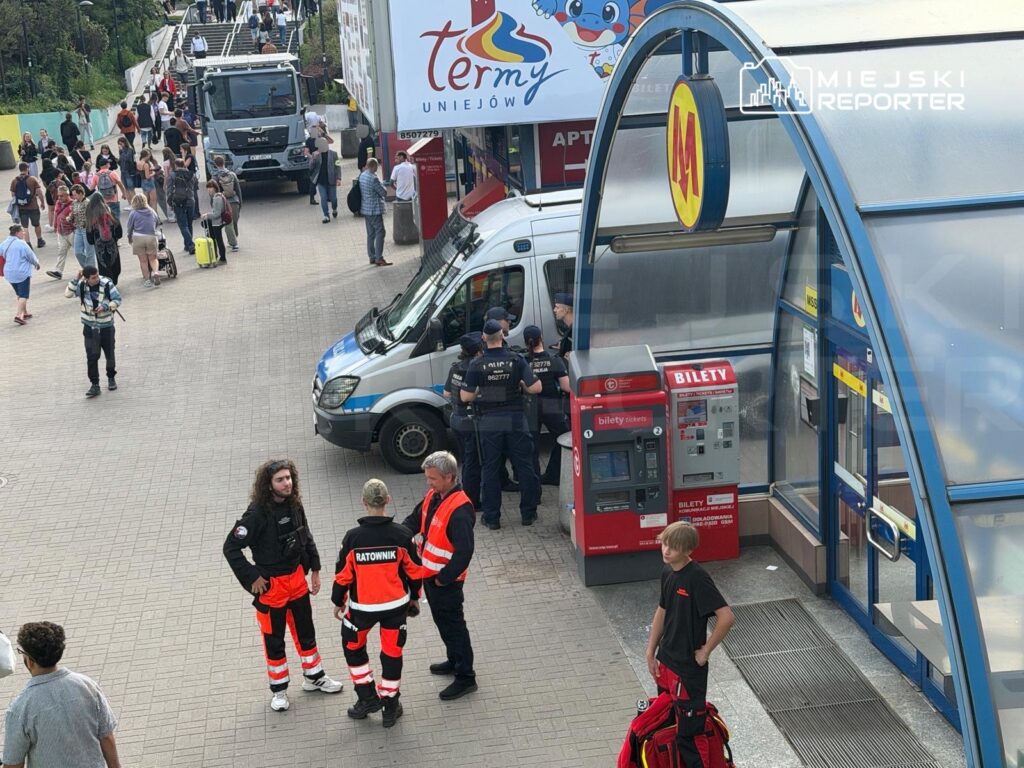 Grupa ratowników w odblaskowych strojach rozmawia w pobliżu stacji, obok radiowozu i automatu biletowego.