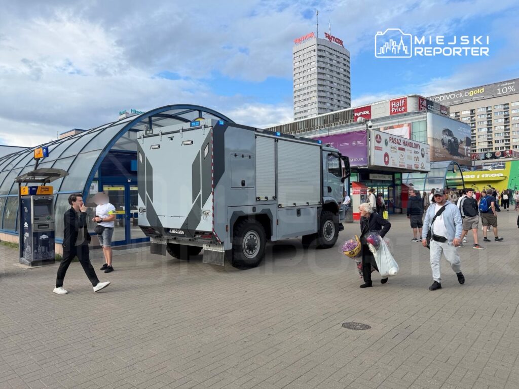 W centrum miasta, obok przeszklonego budynku, stoi pojazd policyjny, a przechodnie idą w różnych kierunkach.