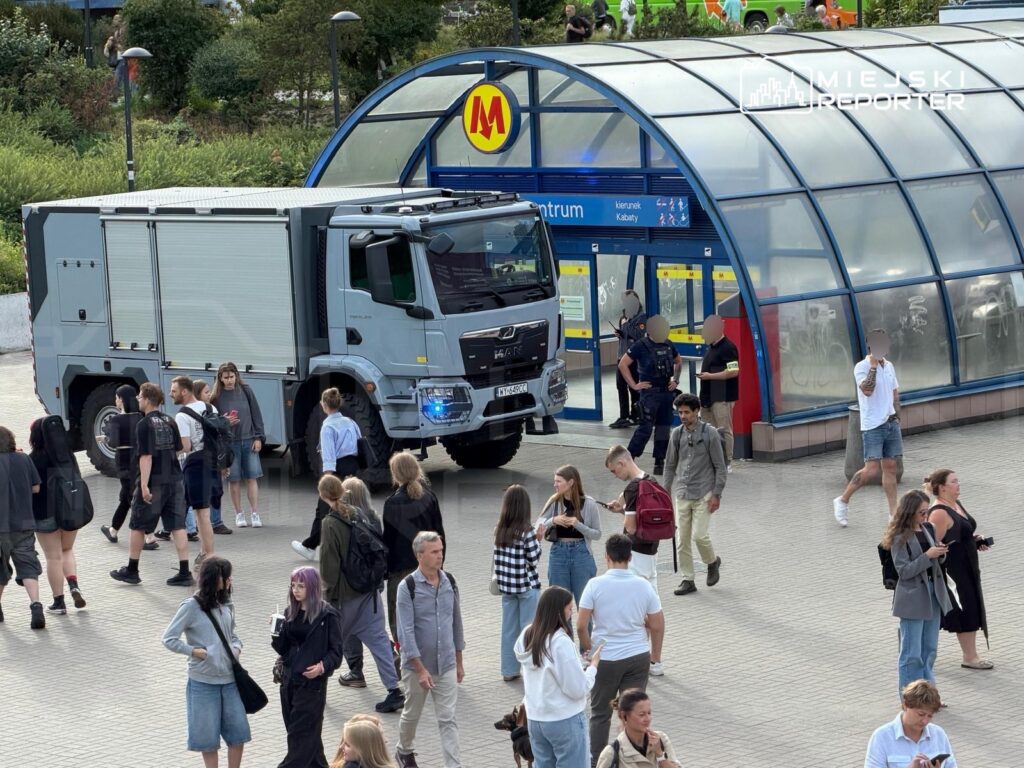Grupa młodych ludzi spaceruje przed stacją metra, obok stoi wóz strażacki z włączonymi sygnałami świetlnymi.