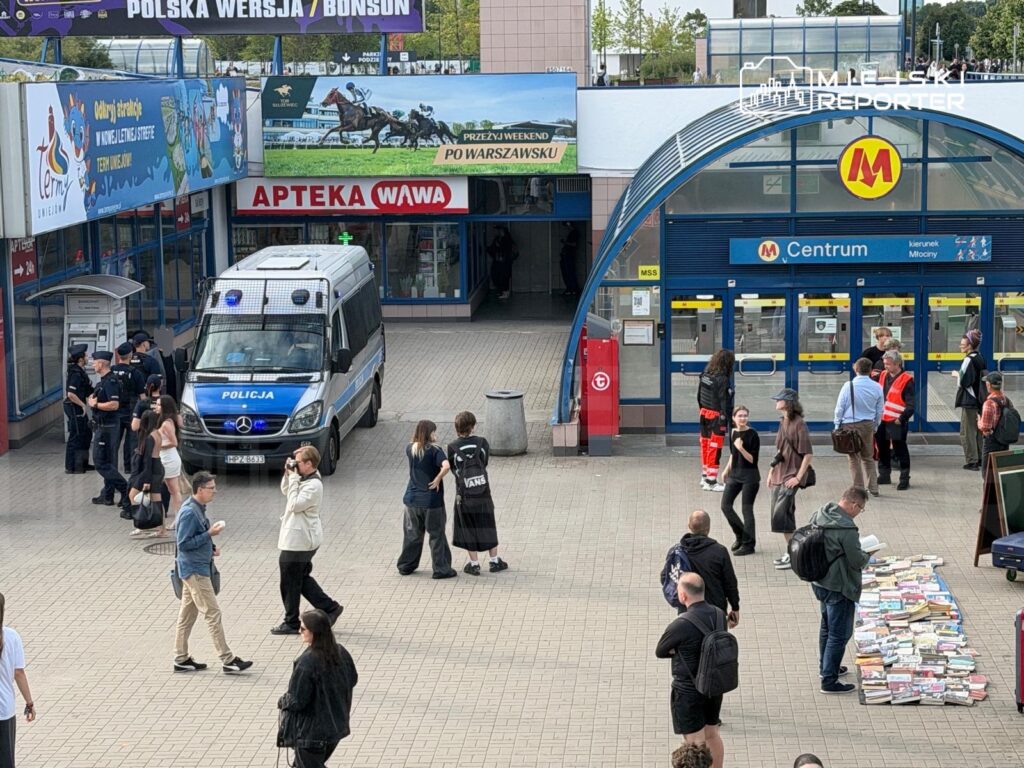 Grupa policjantów przy radiowozie w otoczeniu przechodniów i osób przeglądających książki na chodniku.