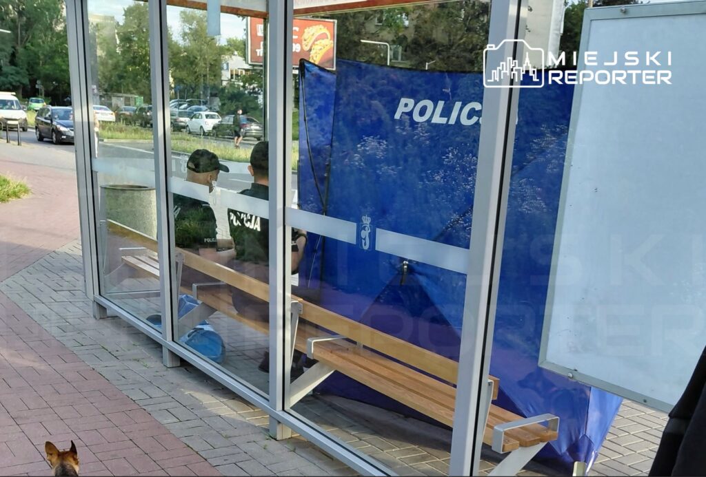 Dwaj policjanci w mundurach siedzą na ławce w przeszklonym punkcie, obok niebieskiej ścianki z napisem "POLICJA".