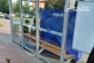 Dwaj policjanci w mundurach siedzą na ławce w przeszklonym punkcie, obok niebieskiej ścianki z napisem "POLICJA".