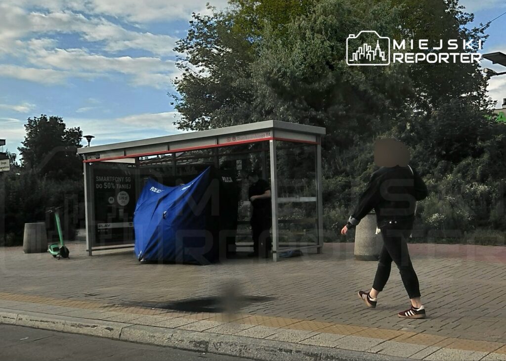 Kobieta w czarnej kurtce spacerująca obok przystanku autobusowego, obok niebieskiego namiotu.
