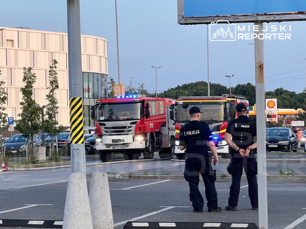 Dwa wozy strażackie z włączonymi sygnałami świetlnymi stoją w pobliżu policjantów, którzy obserwują sytuację na parkingu.