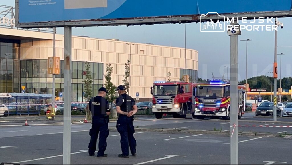 Policjanci rozmawiają obok wozu strażackiego na parkingu przed nowoczesnym budynkiem.