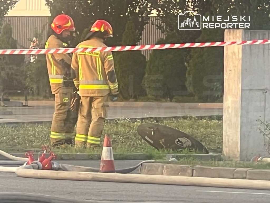 Dwaj strażacy w mundurach z hełmami obserwują miejsce zdarzenia, w tle widoczne węże strażackie i barierki.