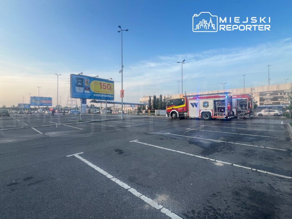 W tle parkingu przed dużym centrum handlowym stoi wóz strażacki z włączonymi sygnałami świetlnymi.
