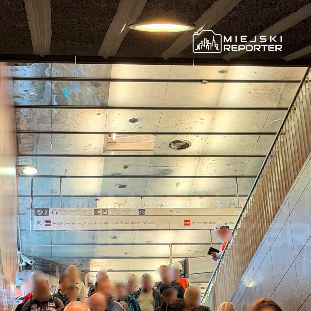 Grupa pasażerów wchodzi po schodach w nowoczesnym obiekcie komunikacyjnym, a nad nimi widoczne są oznaczenia kierunkowe.
