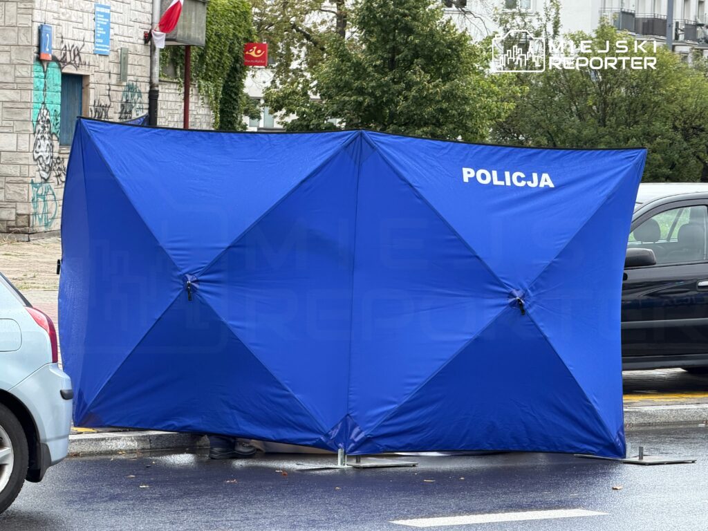 Niebieski namiot policyjny rozstawiony na ulicy, w tle widoczne pojazdy i graffiti na ścianie budynku.