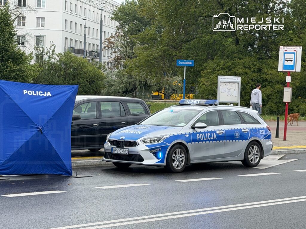 Policyjny radiowóz z niebieską flagą zabezpieczającą miejsce zdarzenia na ulicy, obok przystanku autobusowego.