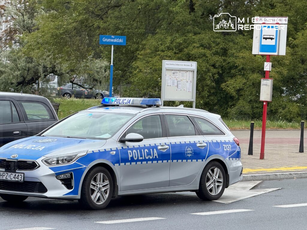 Policyjny samochód na Plac Grunwaldzki z włączonymi sygnałami świetlnymi, obok przystanku autobusowego.