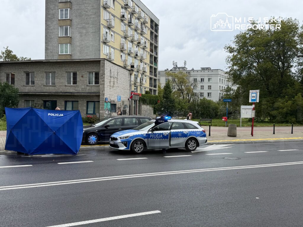 Policyjny radiowóz z włączonymi sygnałami świetlnymi stoi obok niebieskiego namiotu z napisem „POLICJA” na ulicy, w tle budynek mieszkalny.