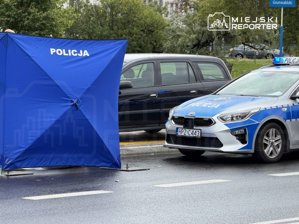 Niebieski namiot policyjny ustawiony na ulicy obok radiowozu z włączonymi sygnałami świetlnymi.