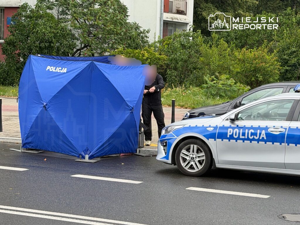 Policjant w czarnym ubiorze prowadzi czynności dochodzeniowe przy niebieskim namiocie z napisem "POLICJA" na ulicy.