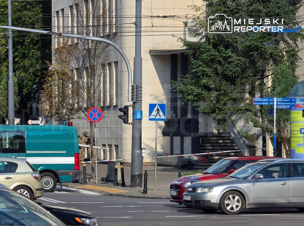 Zielony pojazd służbowy zaparkowany przy budynku, w tle żołnierze stojący za taśmą zabezpieczającą.