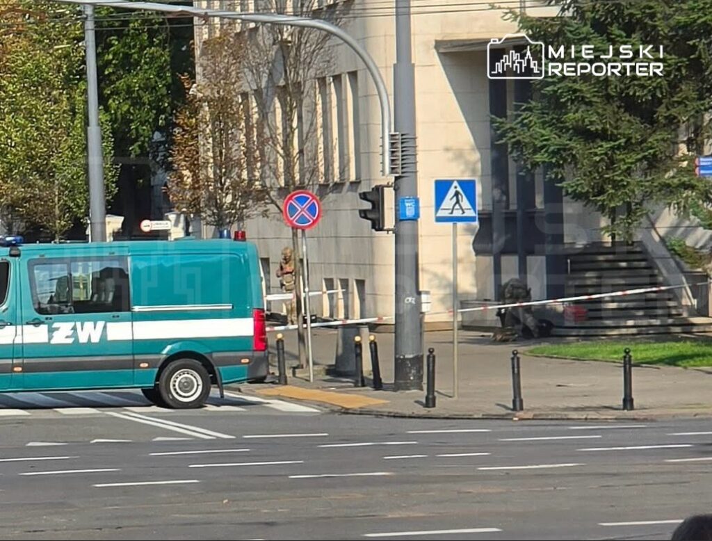 Zielony pojazd służbowy z napisem „ZW” zaparkowany na skrzyżowaniu, w tle widoczna budowla z przeszkloną fasadą.