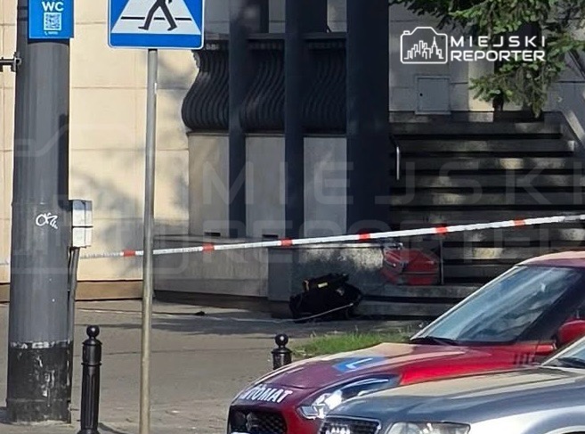 Policyjna furgonetka stoi na chodniku obok oznaczenia przejścia dla pieszych, a w tle widoczny jest ogrodzony teren z torbą.