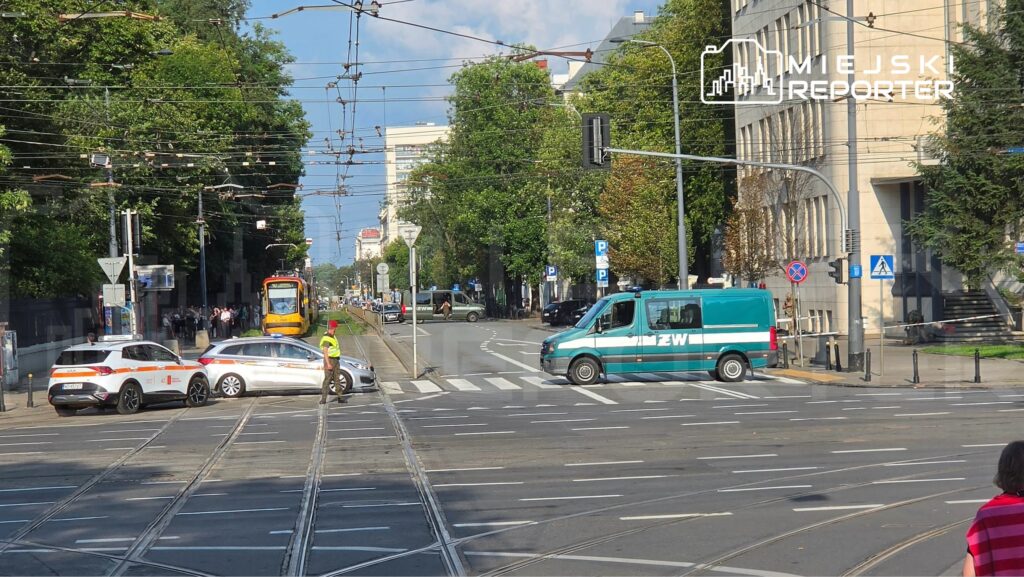 Na skrzyżowaniu ulic stoją dwa samochody służbowe, jeden z nich blokuje torowisko, a obok pracuje strażnik.