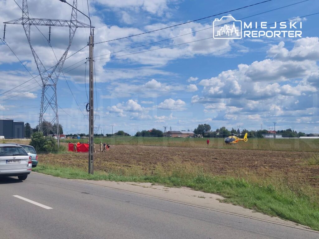 Na polu lądował śmigłowiec ratunkowy, a obok grupka osób w mundurach ustawia czerwoną osłonę.