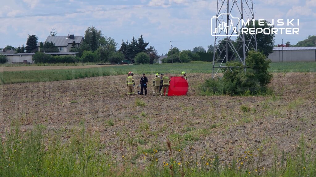 Grupa strażaków i policjantów prowadzi działania na polu, obok czerwonego namiotu ratunkowego. W tle widać domy i wieżę energetyczną.