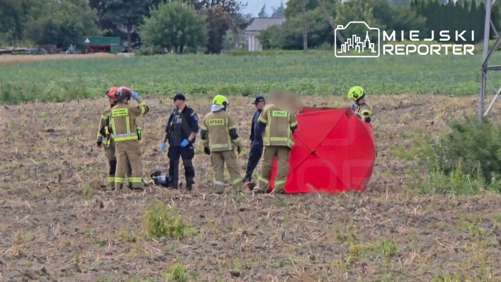 Grupa strażaków i policjantów w specjalistycznym sprzęcie pracuje na polu, obok czerwonego namiotu.