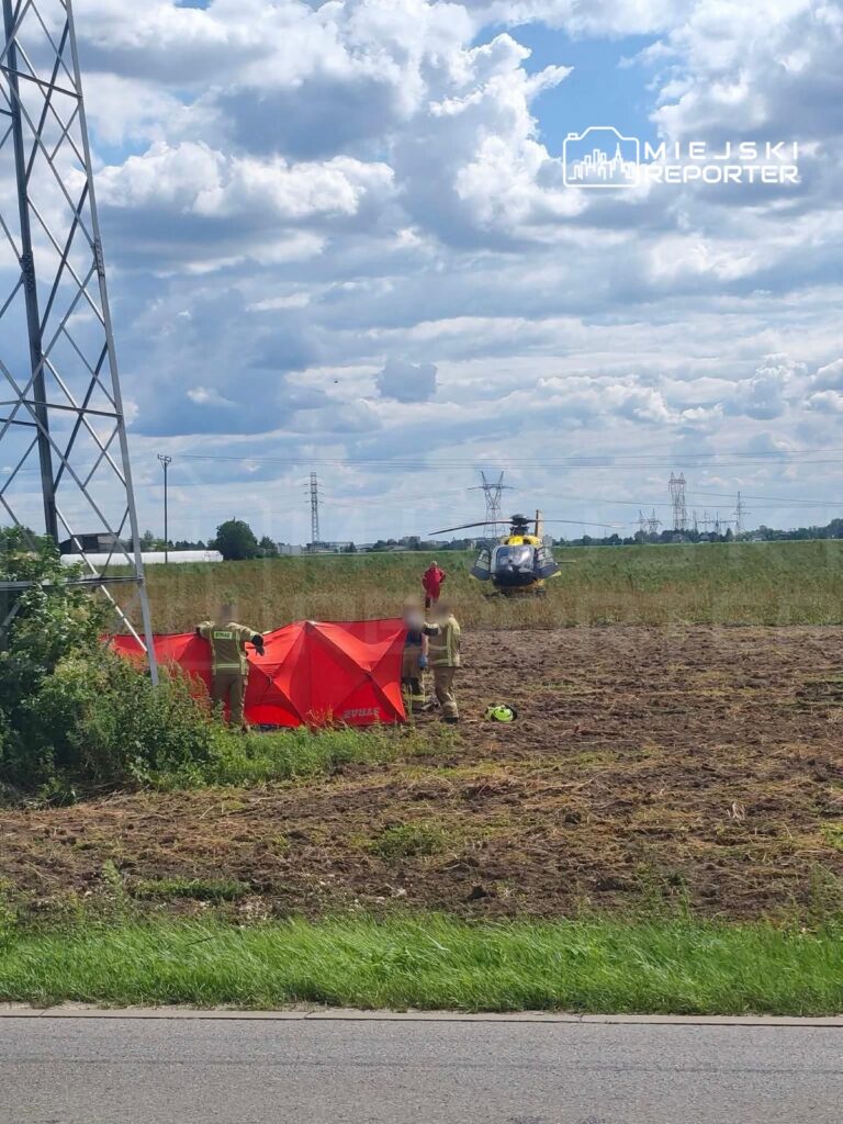 Grupa strażaków ustawia czerwoną osłonę na polu, obok ląduje helikopter ratunkowy. W tle widoczne chmury.