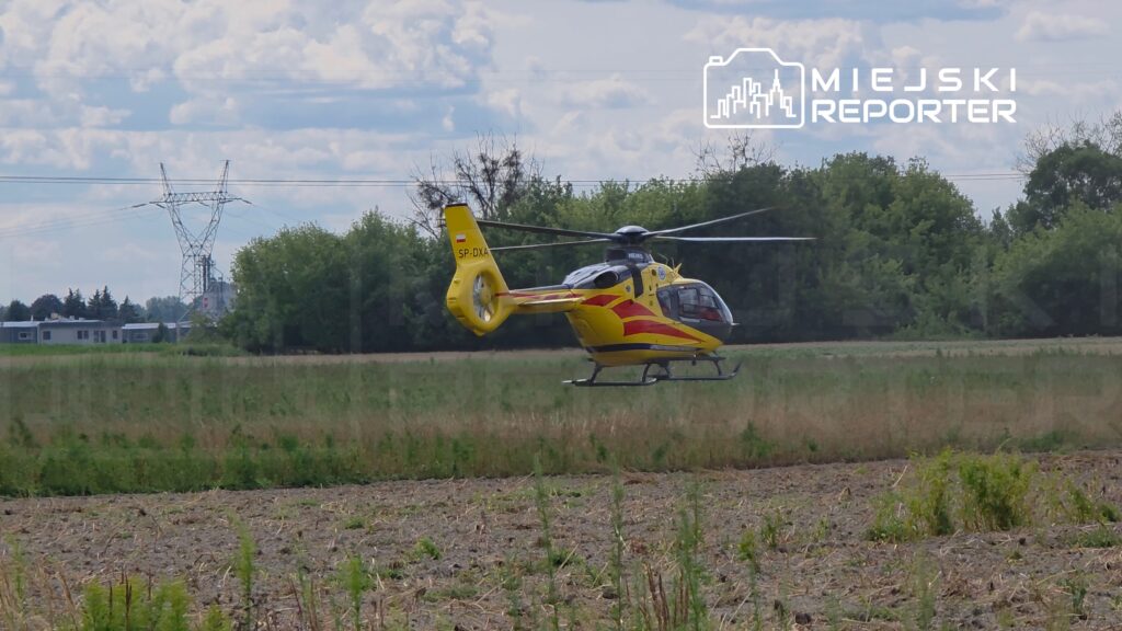 Żółty helikopter unoszący się nad polem, w tle widoczne drzewa i linie energetyczne.