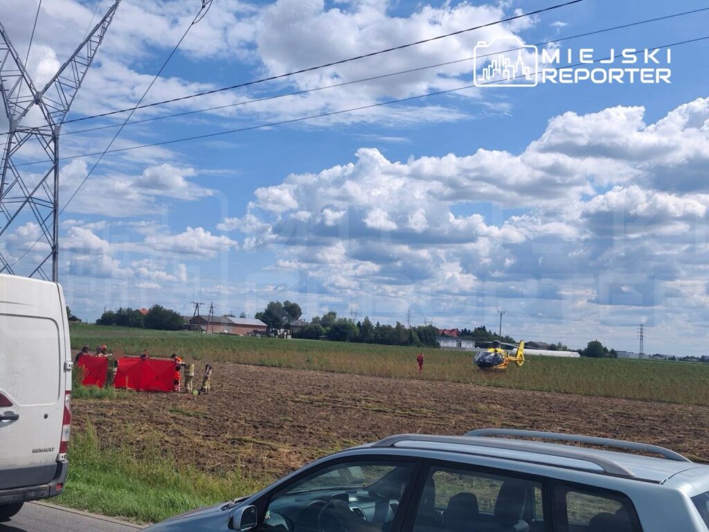 Grupa ratowników w czerwonych strojach pracuje w polu, obok lądowego śmigłowca ratunkowego, na tle błękitnego nieba.