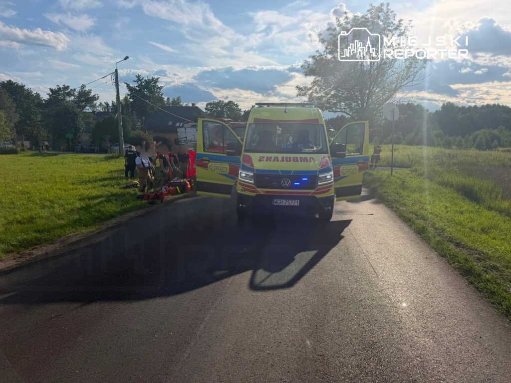Ambulans z otwartymi drzwiami zaparkowany na drodze, obok grupka ratowników udzielających pomocy poszkodowanemu.