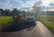 Ambulans z otwartymi drzwiami zaparkowany na drodze, obok grupka ratowników udzielających pomocy poszkodowanemu.
