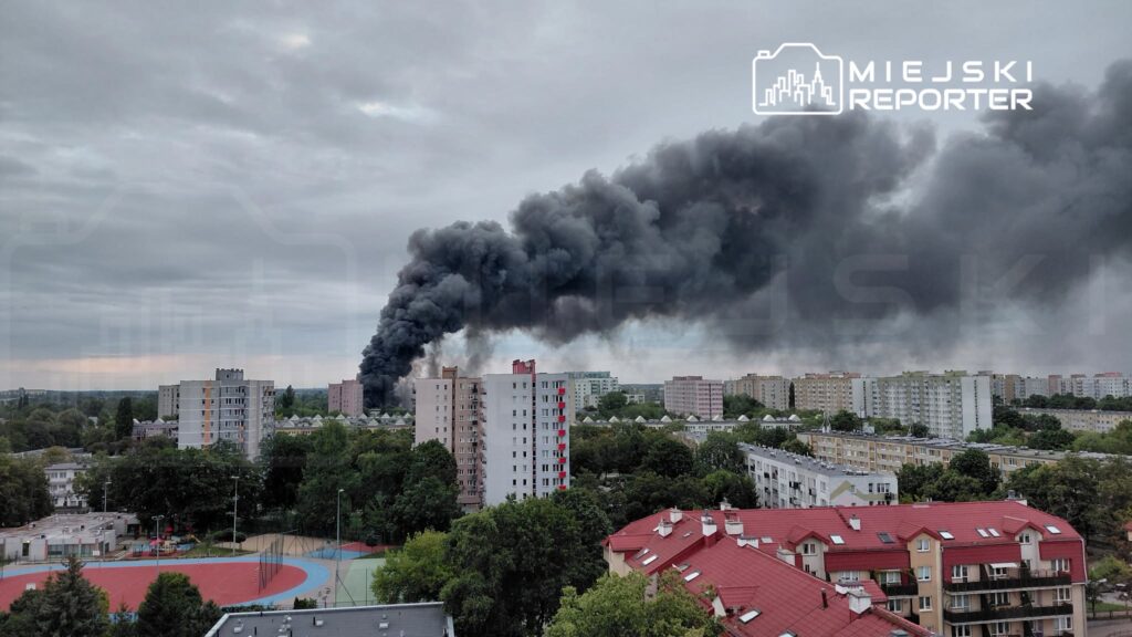 Gęsty czarny dym unoszący się nad zabudowaniami mieszkalnymi w mieście, w tle widoczne bloki i chmury.