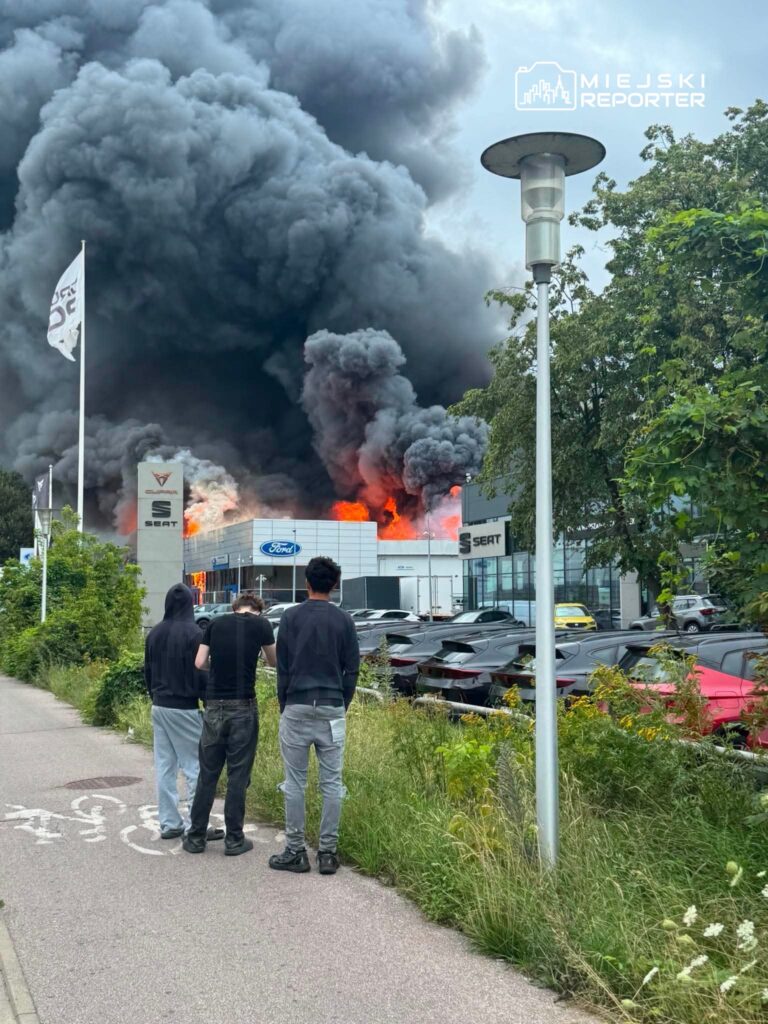 Trzech mężczyzn obserwuje gęsty czarny dym i płomienie wydobywające się z budynku, w tle samochody na parkingu.