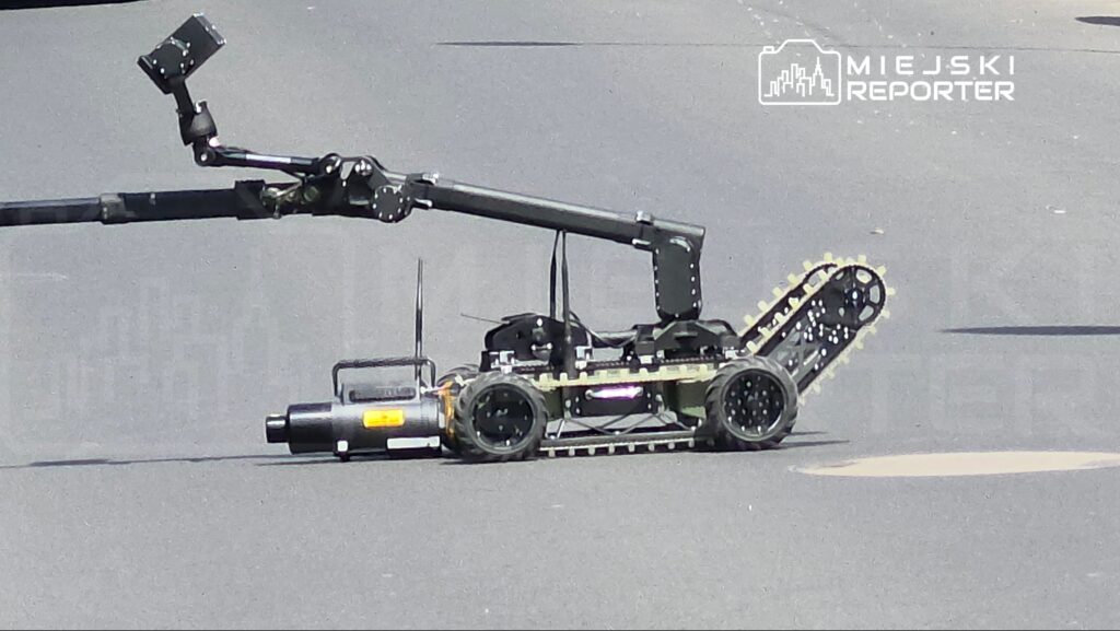 Robot z gąsienicami i wysięgnikiem bada nawierzchnię drogi, wyposażony w kamerę oraz czujniki.