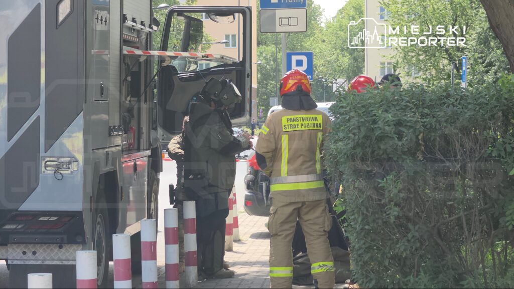 Strażacy w mundurach omawiają działania przy specjalistycznym wozie strażackim na miejskiej ulicy.