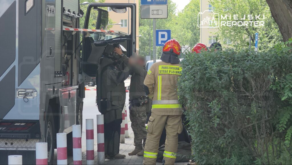 Grupa strażaków w mundurach omawia działania obok specjalistycznego pojazdu strażackiego na miejskiej ulicy.