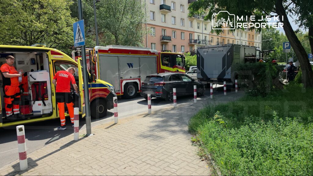 Ratownicy medyczni w pomarańczowych strojach pracują przy ambulansie na miejskiej ulicy, obok strażackiego wozu.