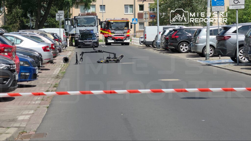 Robot zdalnie sterowany sprawdzający niebezpieczny przedmiot na drodze, otoczonej zaparkowanymi samochodami.